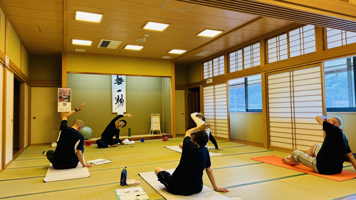 畳の大広間でからだを横に倒すストレッチをする参加者のみなさん